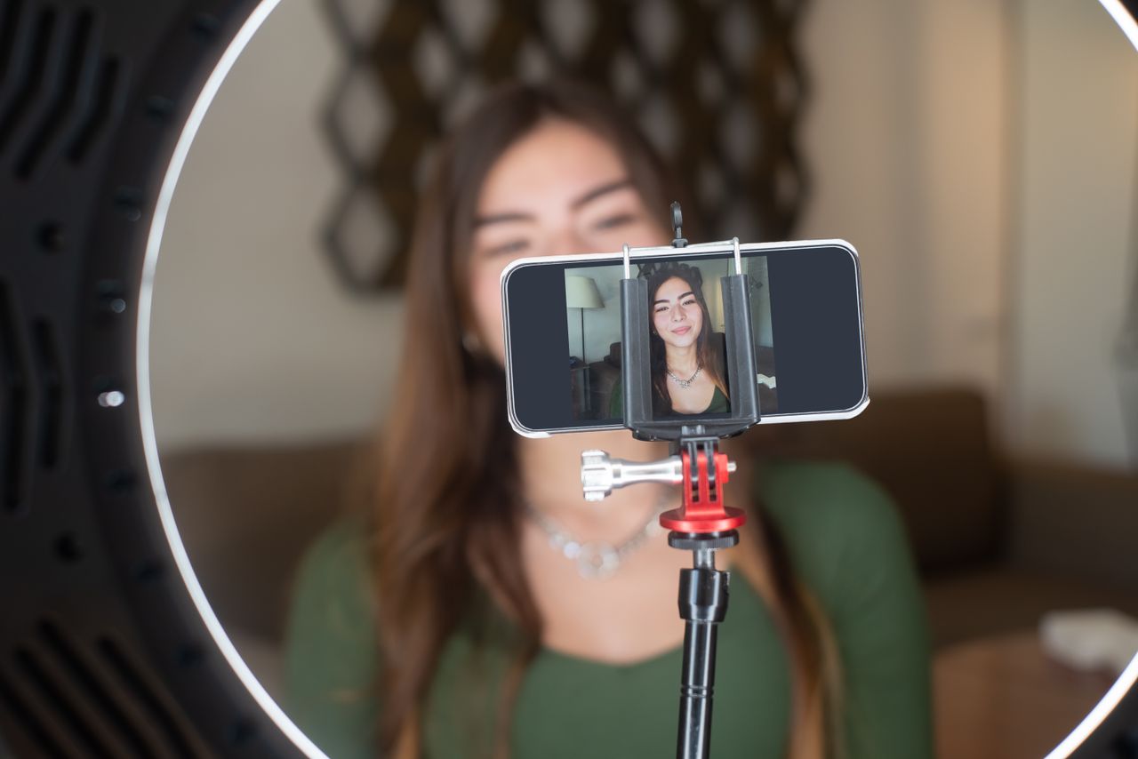 mobile-01 A young woman live streams from home using a smartphone and ring light setup.