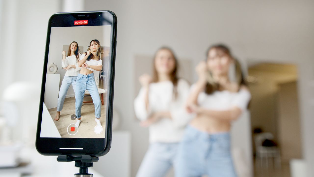 Services-01 Two women dance in front of a smartphone camera, recording content indoors.